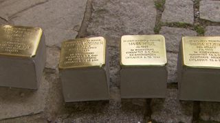 Four gold plaques on concrete blocks, each with names and dates, lie on a ground. Two plaques read 'Maria Holzl' and 'Im Widerstand verhaftet'. Another plaque reads 'Gia Kosssner' and 'Im Widerstand verhaftet'. The fourth plaque is partially visible.