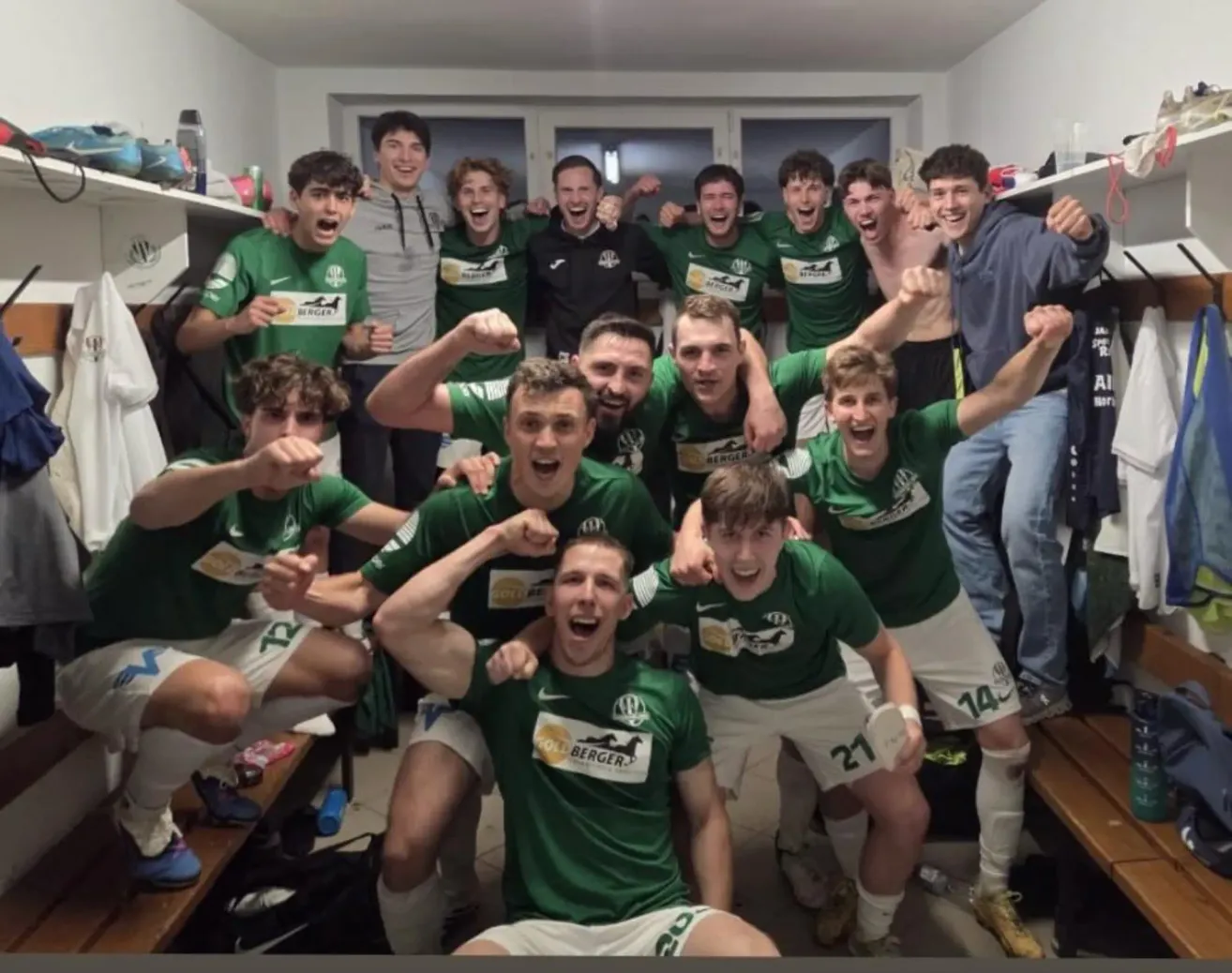 A soccer team in green jerseys poses for a photo in a locker room. They have numbers on their jerseys and appear to be celebrating.