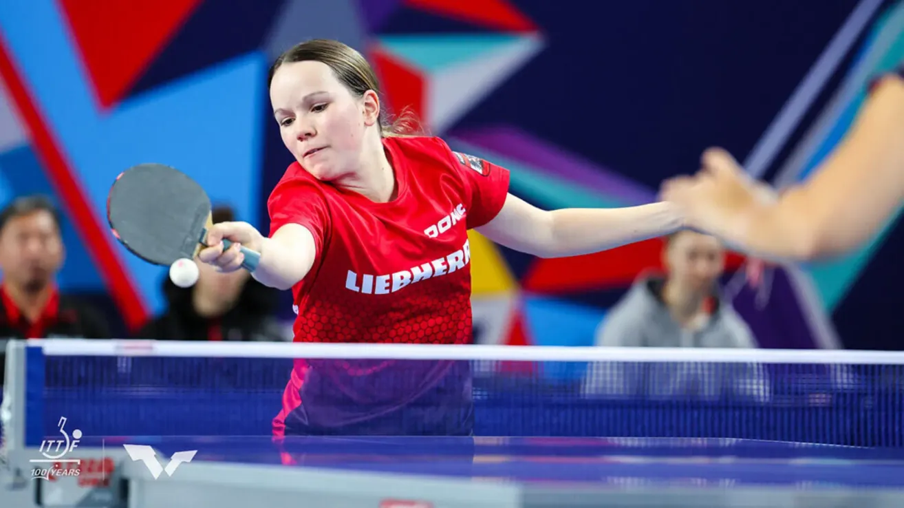 Eine Frau mit einem roten Shirt, auf dem 'DONC' steht, spielt Tischtennis. Sie hält einen Schläger und ist dabei, den Ball zu schlagen.