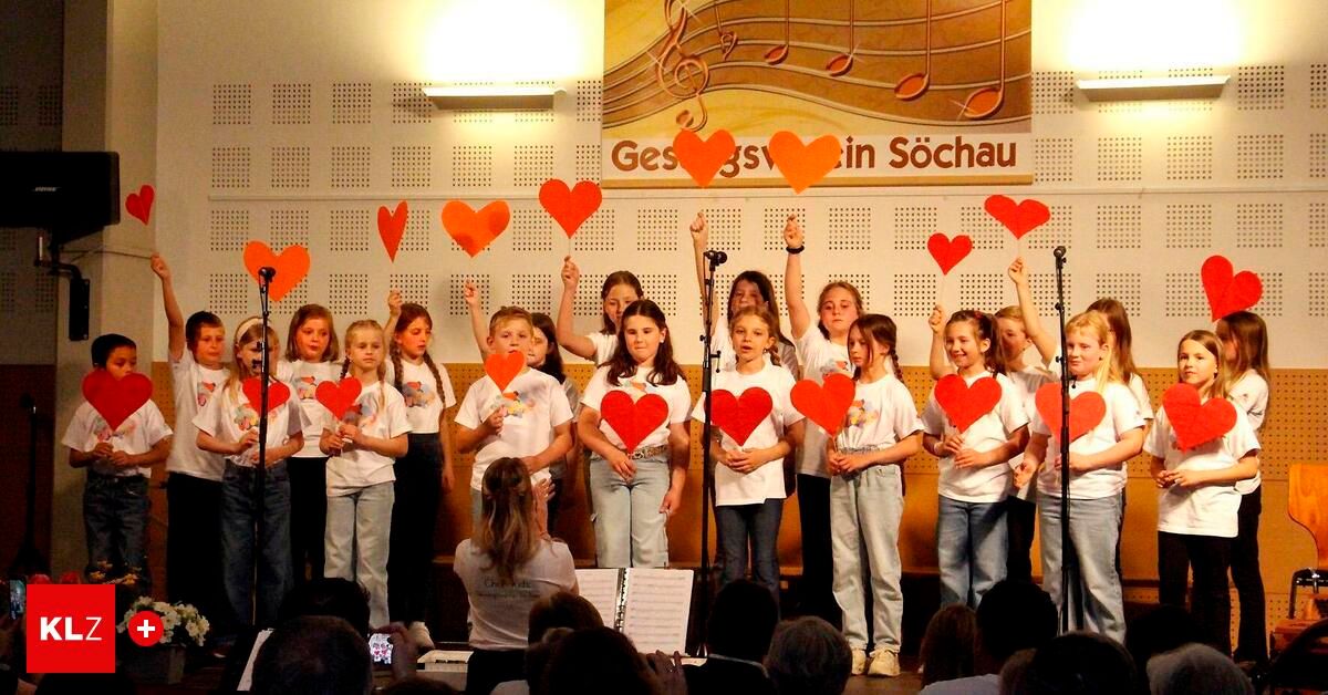 A group of young girls in matching white shirts are performing on stage, holding heart-shaped cutouts. A banner behind them says 'Gess in Sochau'. They are in front of a crowd.