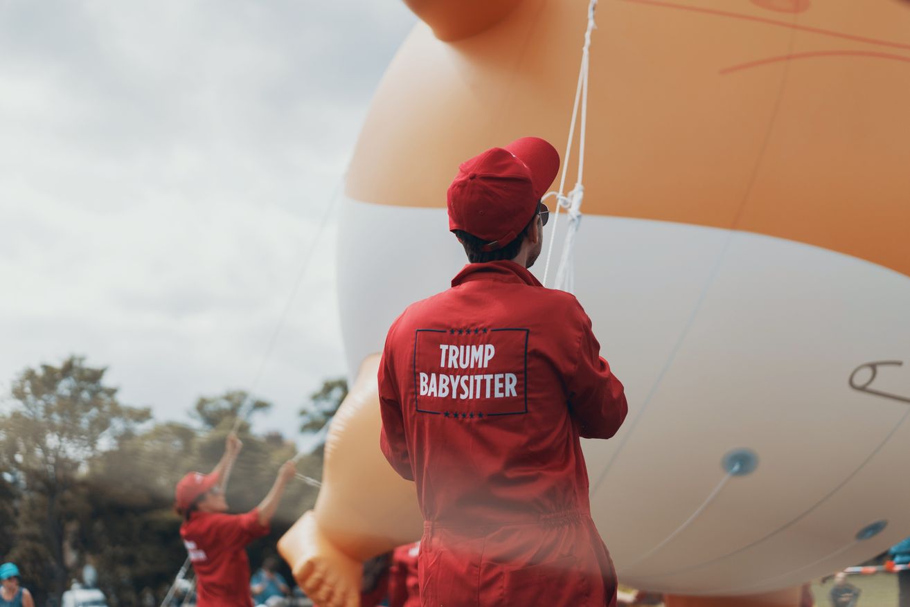 A man in a red jumpsuit and hat holds a rope attached to a giant balloon with the words Trump Babysitter.