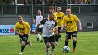 A soccer game in progress. Players in yellow and white jerseys are running, one holding the ball. The field has green grass, and spectators are watching from behind a fence.