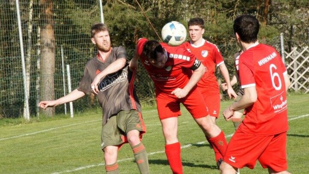 Fußballspieler in roten Uniformen versuchen, den Ball zu köpfen, während ein Spieler in einem grauen Shirt verteidigt.