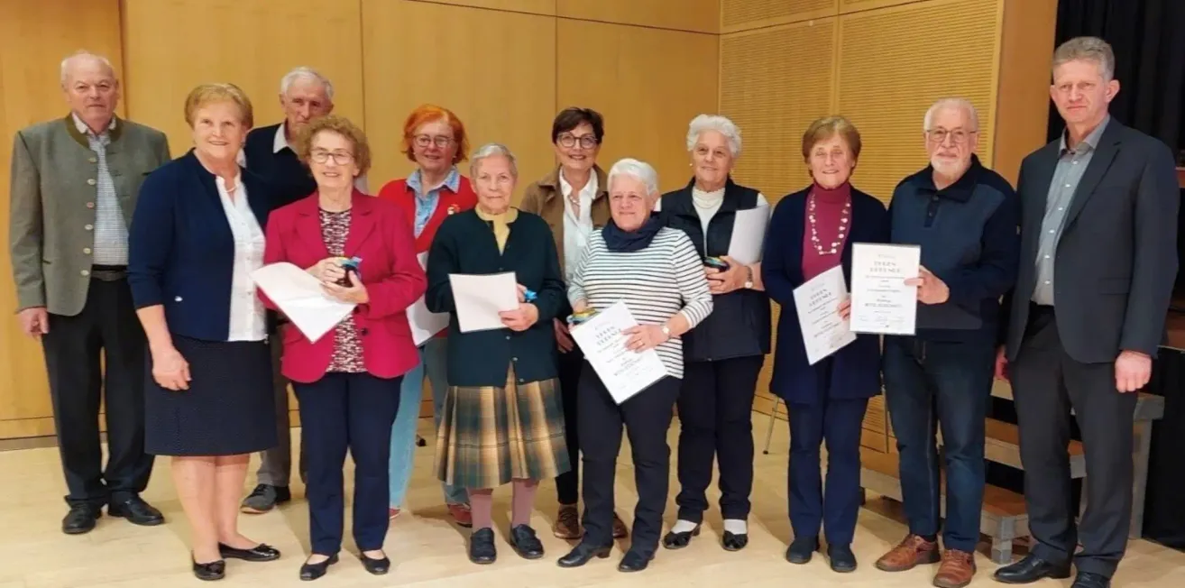 Eine Gruppe älterer Frauen steht zusammen, hält Urkunden und lächelt. Sie tragen formelle Kleidung, und eine hält ein Buch. Sie befinden sich in einem Raum mit einer Holzwand.