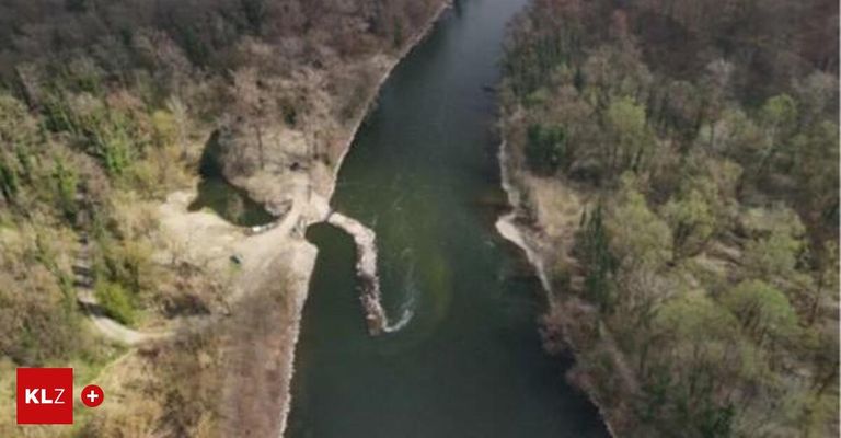 An aerial view of a river flowing through a forest with trees on both sides. The riverbanks are mostly bare, with some small plants and bushes.