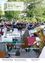 Ein Orchester spielt im Park vor vielen Zuschauern. Der Dirigent steht in der Mitte, und die Band spielt Tuba, Geige und Trompete. Ein Banner lautet 'Unser Ort feiert 35. Jubiläum'.