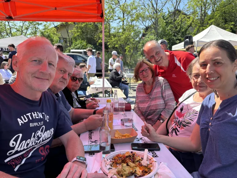 A group of people sitting at a table with food, drinks, and cellphones. Behind them, there are more people under a tent with trees in the background.