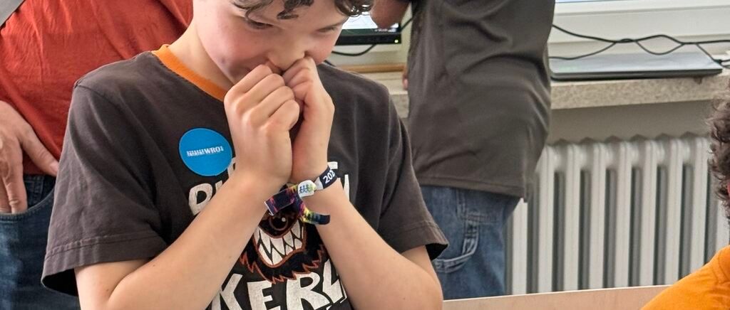 Ein junger Junge mit einem Shirt mit Grafik und dem Jahr 2021 sitzt mit den Händen vor dem Mund. Dahinter steht eine Person in Jeans und einem Shirt neben einem Heizkörper.