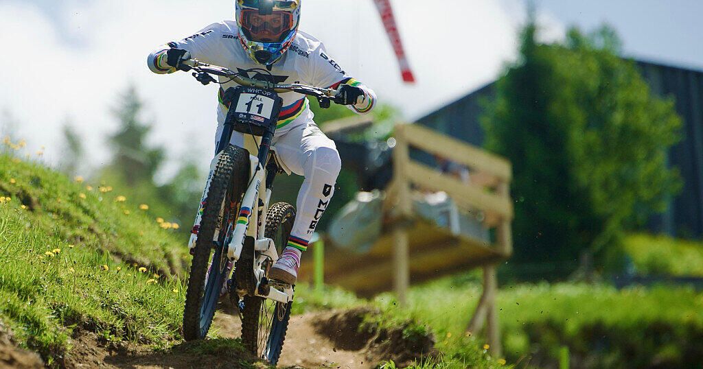 A cyclist wearing a white uniform and a helmet rides down a steep slope. In the background, a wooden structure is visible.