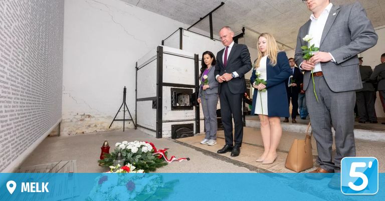 Drei Personen stehen vor einem Denkmal und halten Blumen. Dahinter steht ein weißer Ofen, und eine Frau steht links vom Mann.
