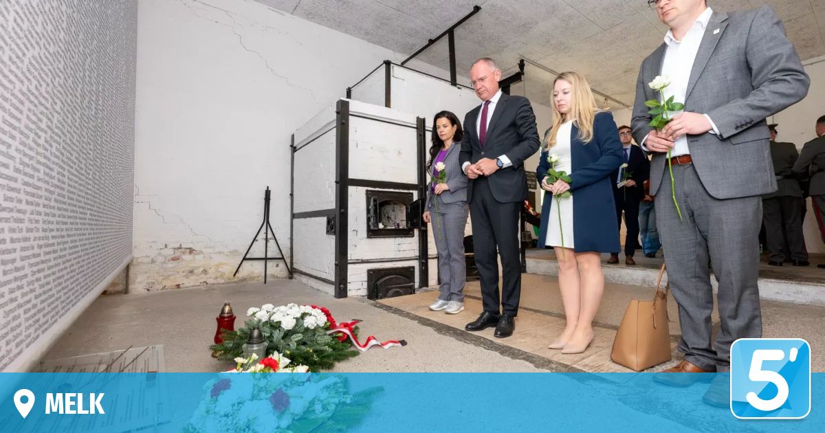 Drei Personen stehen vor einem Denkmal und halten Blumen. Dahinter steht ein weißer Ofen, und eine Frau steht links vom Mann.
