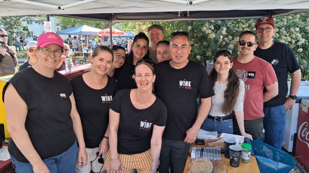 Eine Gruppe von Menschen in schwarzen WINK-T-Shirts steht unter einem Zelt und lächelt für ein Foto. Im Hintergrund sind weitere Personen und Zelte zu sehen.