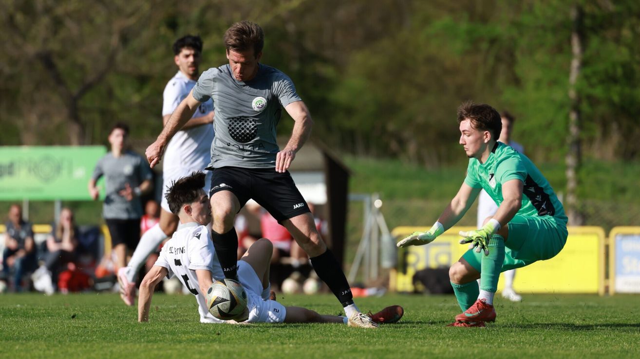 Ein Fußballspieler tritt den Ball. Zwei Spieler versuchen, den Ball zu blockieren. Sie tragen unterschiedlich farbige Uniformen.