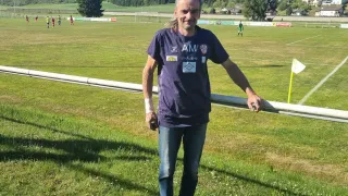 A man is standing on a soccer field. He is smiling and wearing a purple shirt with a logo. Behind him, there are players playing soccer, and a flag is on the ground.