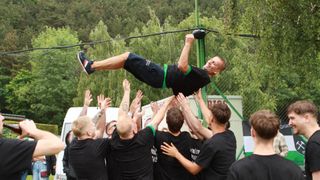 A man in a black shirt is suspended by his hands in the air, supported by several people in black shirts, outdoors near a fence with trees in the background.