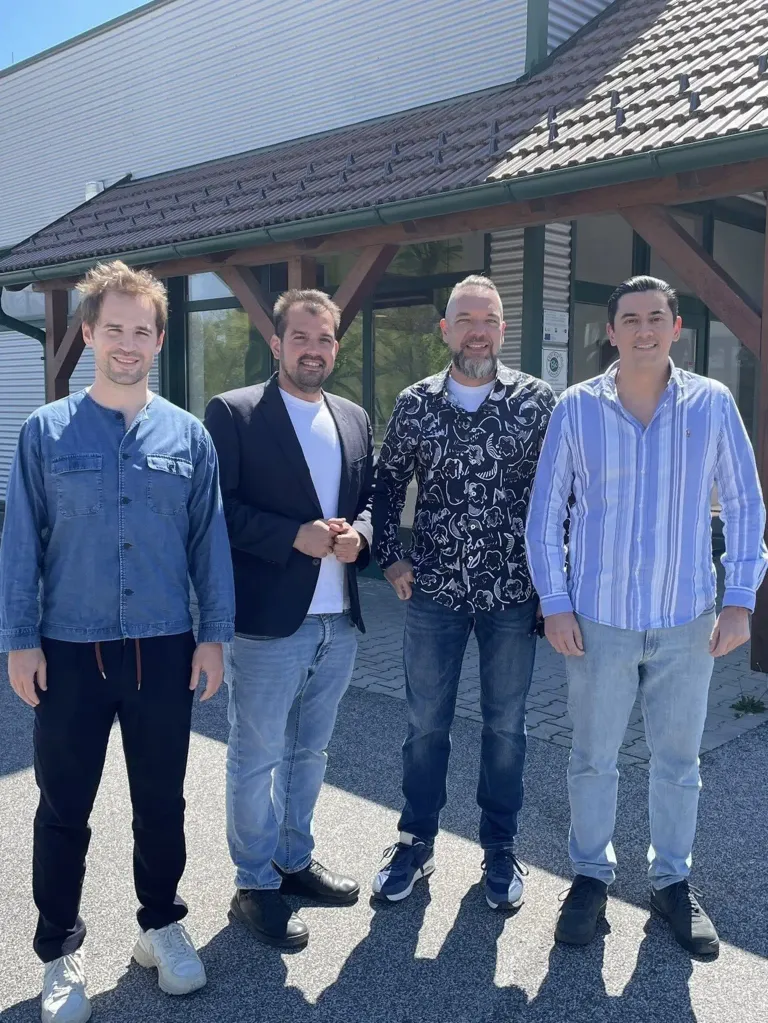 Vier Männer stehen vor einem Gebäude. Sie lächeln und scheinen für ein Foto zu posieren. Ein Mann trägt einen Anzug und einer eine Jeansjacke.