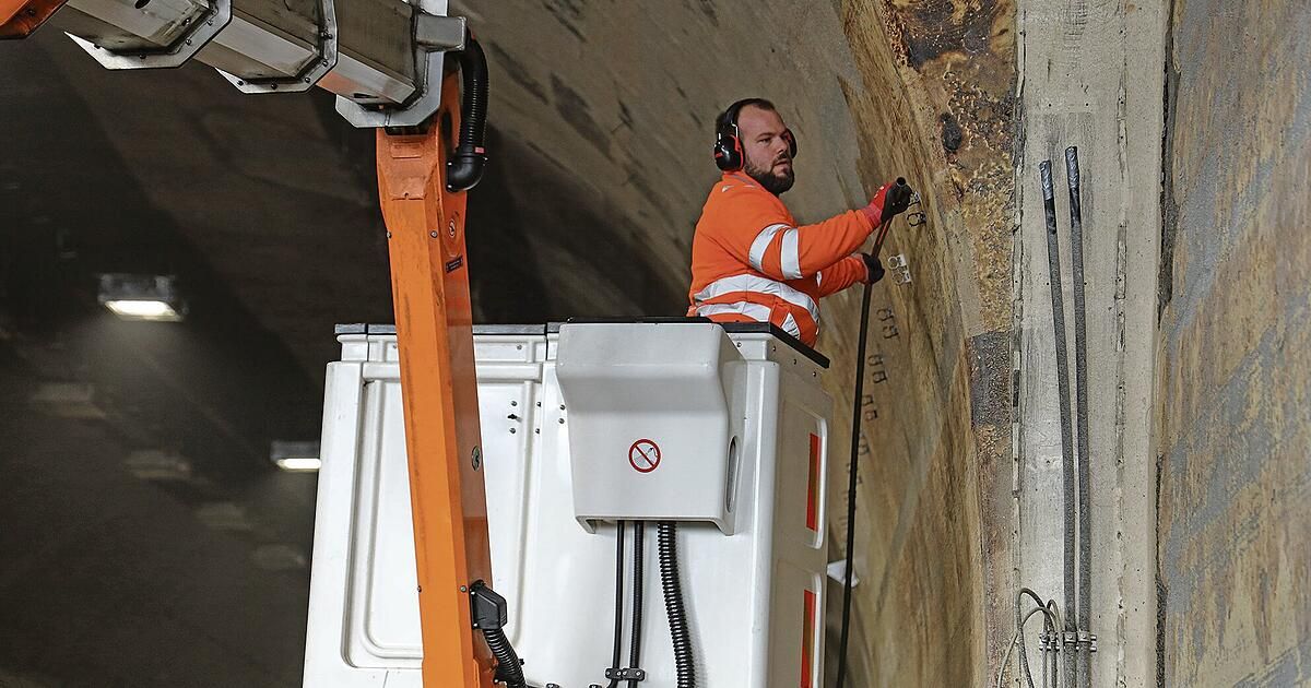 Ein Arbeiter in einem orangefarbenen Sicherheitsanzug reinigt mit einem Schlauch die Seitenwand eines Tunnels. Er steht auf einer weißen Plattform mit einer weißen Maschine und einem roten Rauchverbotsschild.