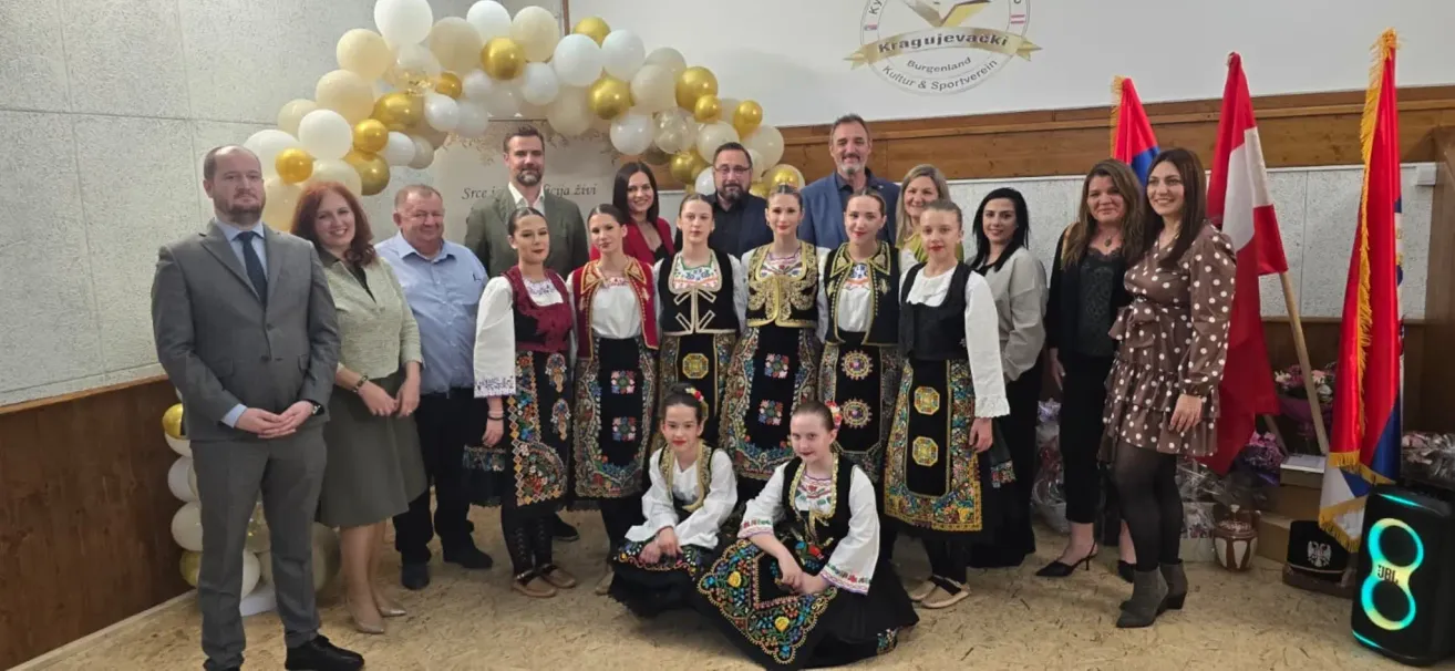 Eine Gruppe von Männern und Frauen in traditionellen Kostümen posiert für ein Foto vor einem Hintergrund mit Luftballons und einem Logo.