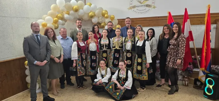 Eine Gruppe von Männern und Frauen in traditionellen Kostümen posiert für ein Foto vor einem Hintergrund mit Luftballons und einem Logo.