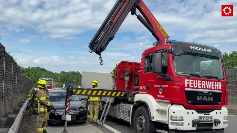 Feuerwehrleute verwenden einen Kran, um ein unter einem Lastwagen festsitzendes Auto zu entfernen. Der Kran ist ausgeklappt und hat einen Haken angebracht. Die Szene spielt im Freien.