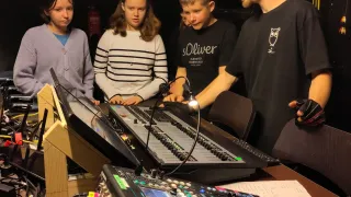 A group of children and a man are in a studio with audio equipment. The man is teaching them about mixing and they are listening attentively.