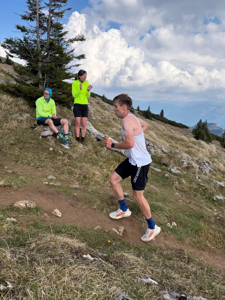 Ein Mann läuft auf einem Pfad in den Bergen. Zwei Personen sitzen hinter ihm auf dem Grashang. Eine Frau steht und ein Mann sitzt auf dem Felsen. Sie tragen alle Sportbekleidung. Der Himmel ist bewölkt.