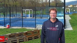 A man wearing a navy blue hoodie with World Padel Tour logo stands in front of an enclosed tennis court. The court is blue with a net and surrounded by wooden pallets.