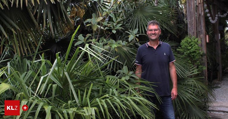 A man stands in a lush tropical garden, wearing a dark blue polo shirt and jeans. He smiles and poses for a photo.