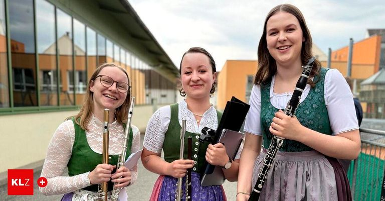 Drei junge Frauen in traditioneller Kleidung halten Musikinstrumente und lächeln vor einem Gebäude. Die mittlere Frau hält eine Flöte, die links eine Klarinette und die rechts eine Flöte.