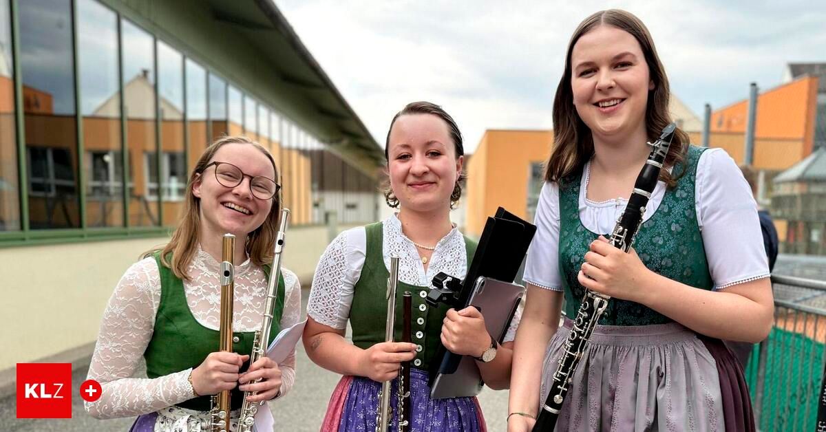 Drei junge Frauen in traditioneller Kleidung halten Musikinstrumente und lächeln vor einem Gebäude. Die mittlere Frau hält eine Flöte, die links eine Klarinette und die rechts eine Flöte.