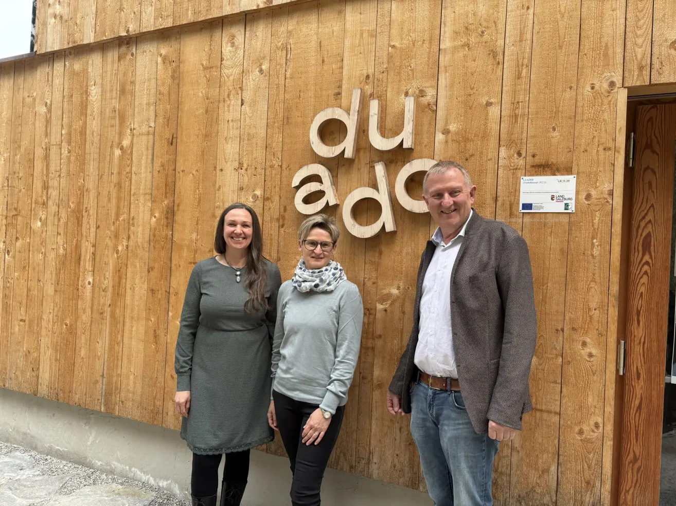 Drei Personen stehen vor einer Holzwand mit den Buchstaben 'du ad'. Die Frau links trägt ein graues Kleid, die mittlere eine Schürze und der Mann einen Anzug.