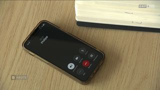 A black smartphone is on a wooden surface with icons for calling, voicemail, and more. The brand name 'Philipp' is displayed on the screen. A notebook with a black cover is partially visible in the background.