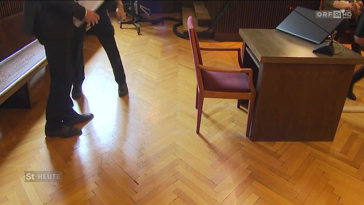 A person is walking on a wooden floor with a red chair and a desk nearby. A staircase and a black hose are also visible.