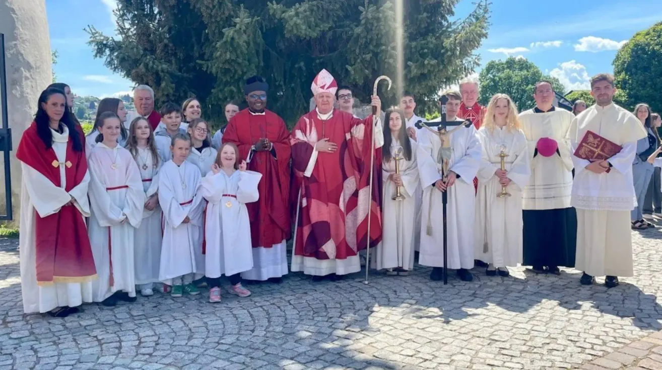 A group of people dressed in white robes stand together with a bishop in red robes. They are holding a cross and a staff. The setting is outdoors with trees and sunlight.