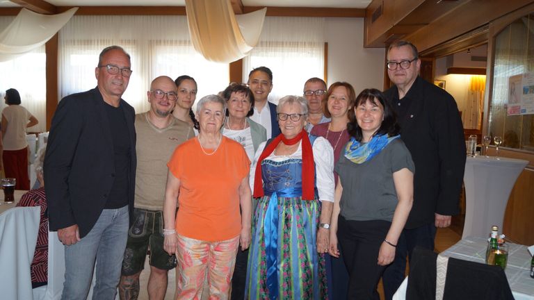 Eine Gruppe von Menschen, sowohl Männer als auch Frauen, stehen und posieren für ein Foto in einem Raum mit weißen Vorhängen und Holzbalken. Sie alle lächeln.