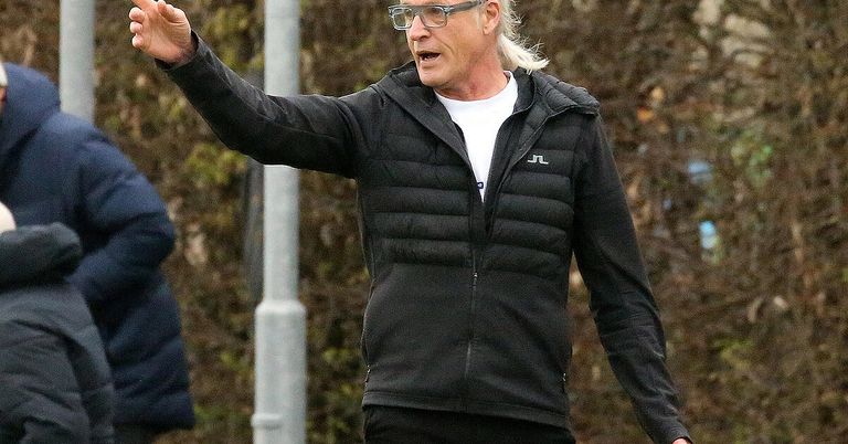 Ein älterer Mann mit langen weißen Haaren und einer Brille trägt eine schwarze Jacke. Er gestikuliert mit der linken Hand, während seine rechte Hand in der Tasche steckt. Er scheint mit jemandem vor ihm zu sprechen.
