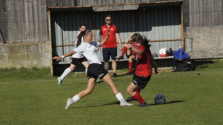 Fußballspieler in weißen und roten Trikots spielen auf einem Feld. Ein Spieler in Weiß tritt den Ball, während ein anderer in Rot versucht, ihn zu blockieren. Ein Mann beobachtet von hinten.