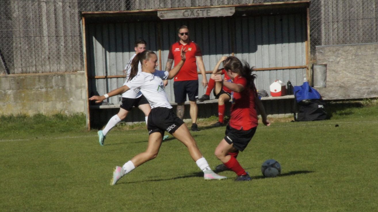Fußballspieler in weißen und roten Trikots spielen auf einem Feld. Ein Spieler in Weiß tritt den Ball, während ein anderer in Rot versucht, ihn zu blockieren. Ein Mann beobachtet von hinten.