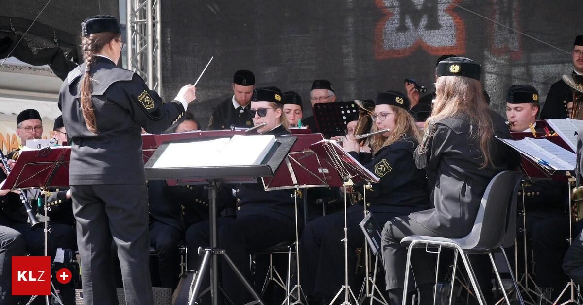 A military band is performing on stage with various instruments, including flutes, trombones, and tubas. The conductor leads the band while seated musicians play their instruments.