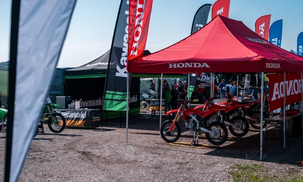 A motorcycle exhibition with a Honda tent displaying several red motorcycles. Banners for Kawasaki and Honda are visible. A Kawasaki motorcycle is parked in the foreground.
