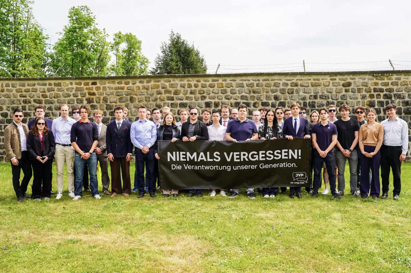 A group of people wearing formal attire stands before a stone wall, holding a black banner with white text that reads 'Niemals Vergessen!' They seem to be posing for a photo.