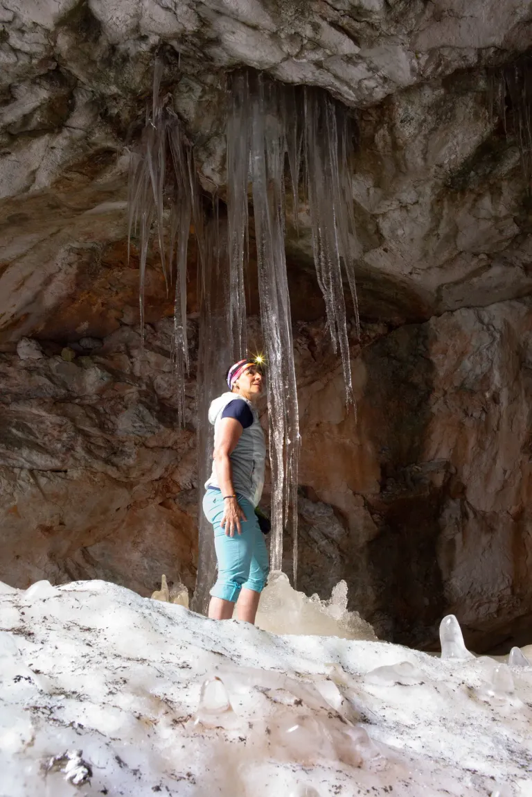 A woman stands in a cave with icicles hanging from the ceiling. She wears a hat and a jacket. She looks up at the icicles.