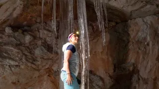 A woman stands in a cave with icicles hanging from the ceiling. She wears a hat and a jacket. She looks up at the icicles.
