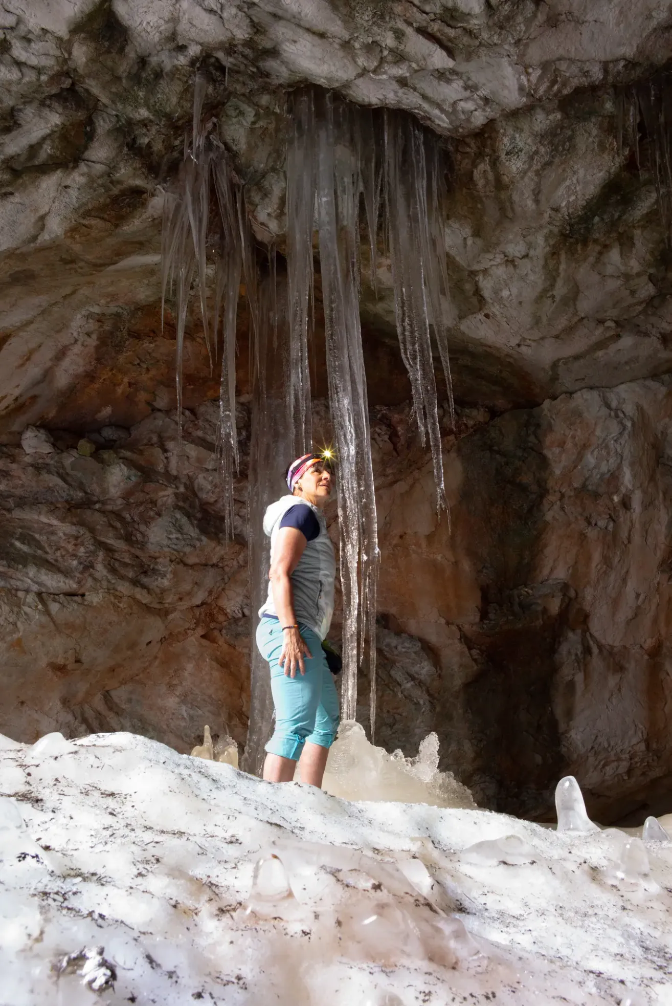 A woman stands in a cave with icicles hanging from the ceiling. She wears a hat and a jacket. She looks up at the icicles.