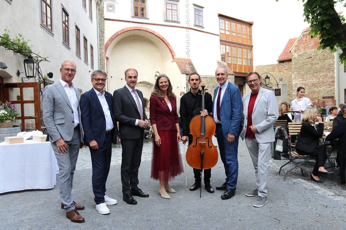 A group of people, including a cellist, stand together in front of an old building.