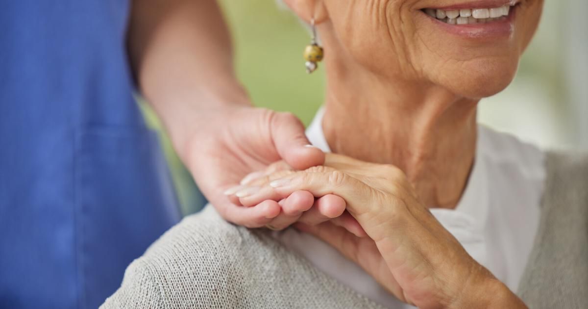 Eine ältere Frau hält die Hand einer Person, möglicherweise einer Krankenschwester, mit einem sanften Lächeln, trägt Ohrringe und einen grauen Pullover.