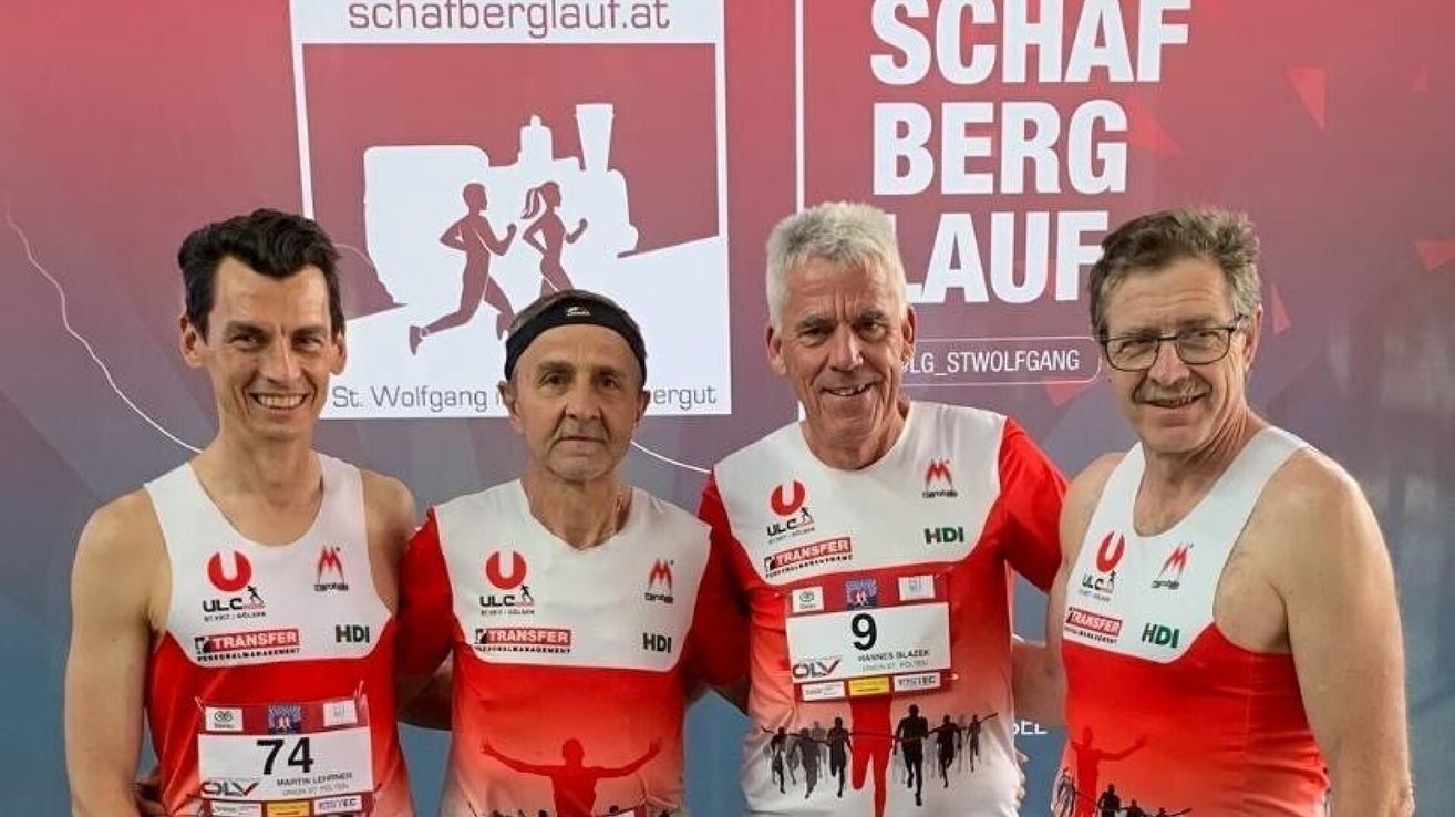 Vier Männer in roten und weißen Trikots stehen beim Schafberglauf-Event zusammen. Ein Banner hinter ihnen hat ein Logo und Text, und ein Mann trägt eine Brille.