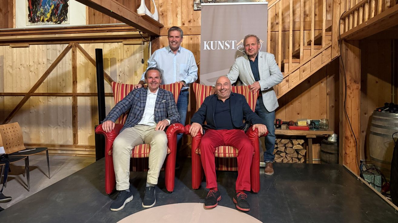 Vier Männer posieren für ein Foto in einem hölzernen Raum, zwei sitzen in roten Stühlen und zwei stehen hinter ihnen, einer hält das Banner mit der Aufschrift 'KUNST'.