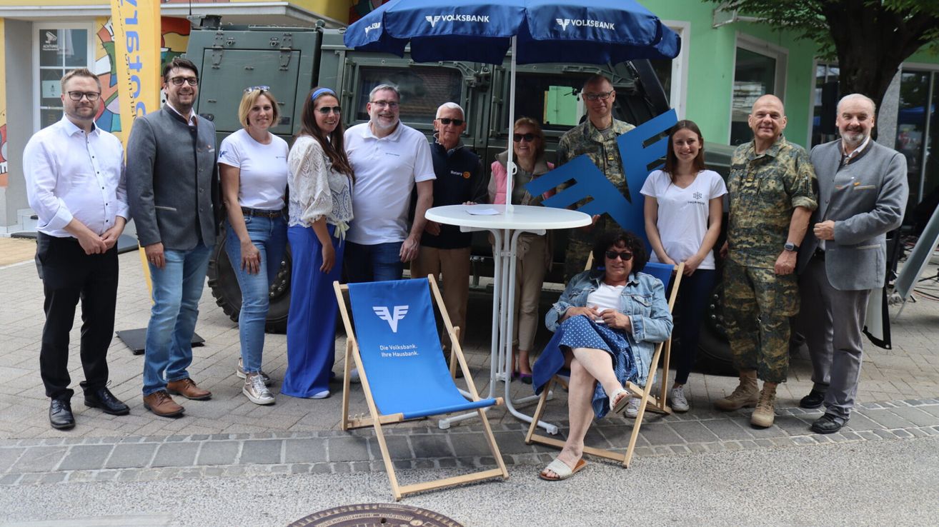 Eine Gruppe von Menschen, darunter eine sitzende Frau und ein Mann in Uniform, posiert für ein Foto vor einem Tisch und einem Schirm. Sie lächeln.