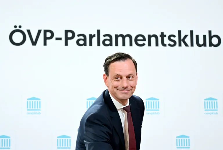 A man in a suit and tie stands in front of a white wall with the word Parlamentsk written on it. He is smiling.